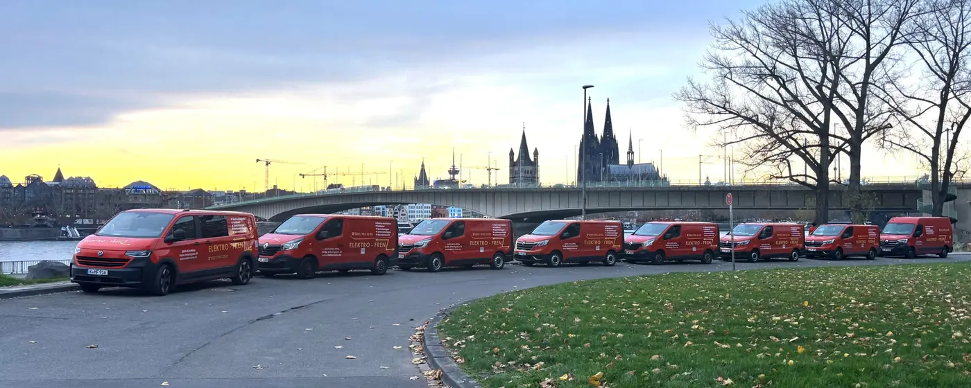 Acht rote Firmenfahrzeuge von Elektro Frenz in einer Reihe am Kölner Rheinufer, im Hintergrund die Brücke und der Kölner Dom